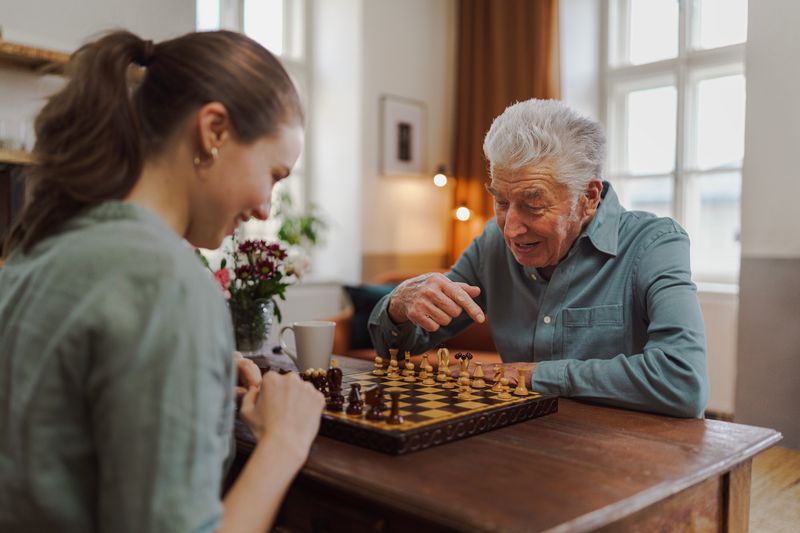 Ein Mädchen sitzt mit einem Senioren am Tisch. Sie spielen Schach und haben Freude dabei.