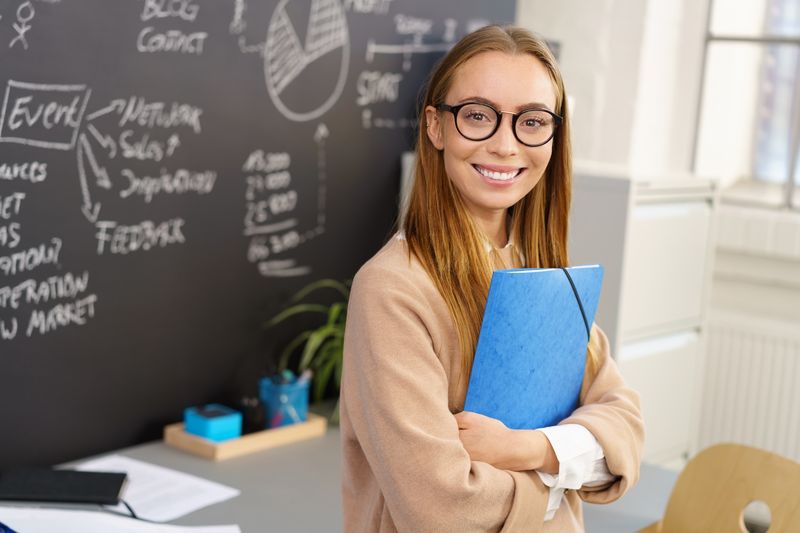 Eine junge Frau mit langen blonden Haaren und Brille, steht vor einem Whiteboard. Sie hält eine Mappe umklammert und lehnt lässig an einem Tisch.