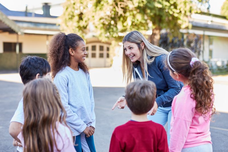 Eine Erzieherin steht mit einer diversen Gruppe von Kindern auf einem Schulhof. Alle lachen ausgelassen und schauen interessiert zur Erzieherin. 