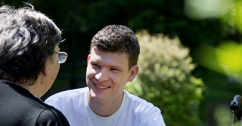 Auszubildender im Bereich Altenpflege unterhält sich lächelnd mit einer Seniorin.