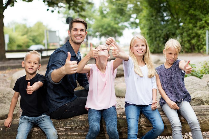 Ein Erzieher ist mit einer Gruppe von Kindern in der Natur. Sie albern herum, ziehen Grimassen und zeigen den Daumen nach oben. 