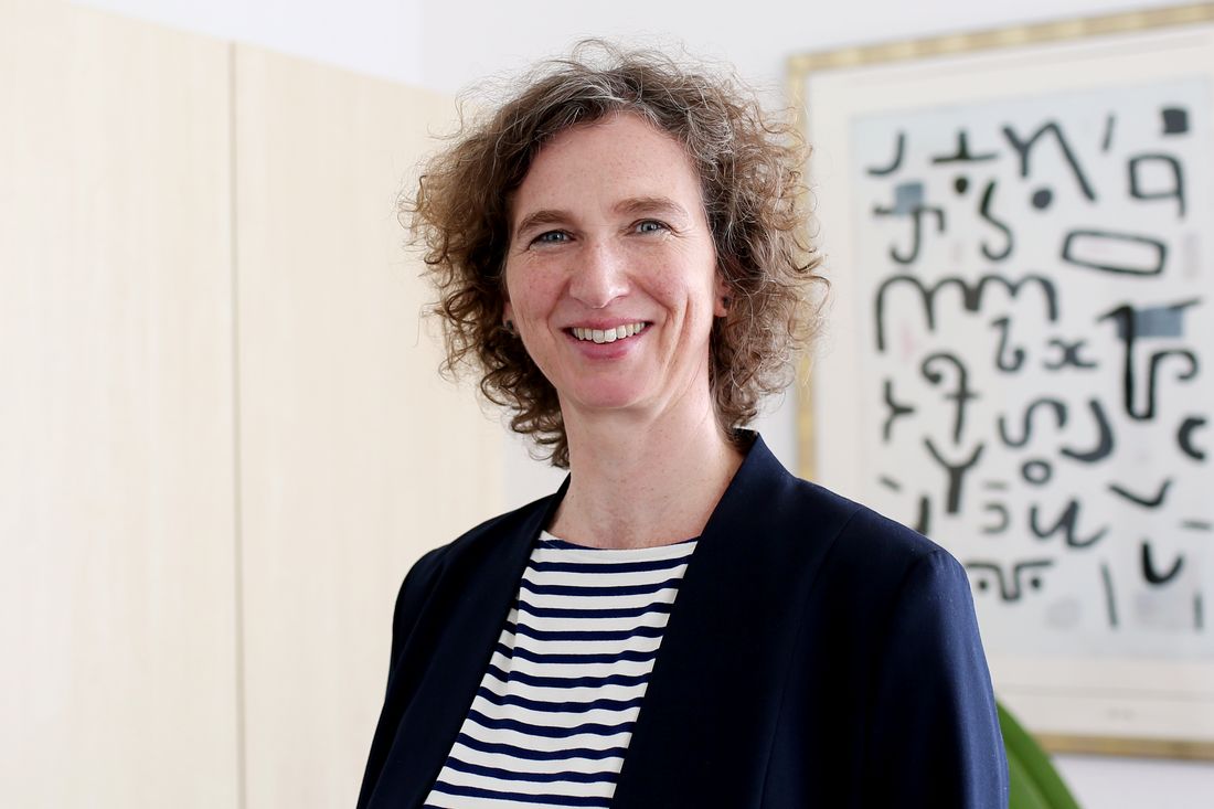 Frau mit Locken steht in einem Büro vor einem Bild und lächelt