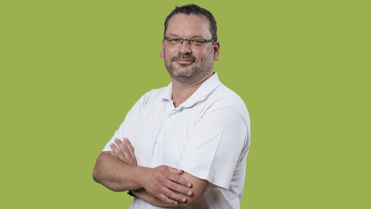 Eine männliche Person mit Bart und Brille trägt ein weißes Hemd. Er steht vor grünem Hintergrund in einer lockeren Pose. Er strahlt Authentizität und Gelassenheit aus.